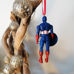 May include: A red, white, and blue Captain America Christmas ornament. The ornament is a plastic figurine of Captain America wearing his iconic costume. He is holding his shield in his left hand and is standing with his feet together. The ornament is hanging from a red ribbon.