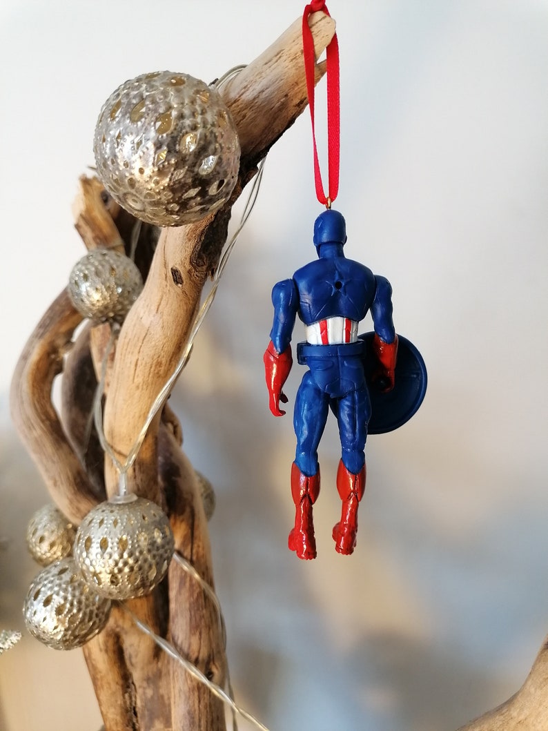 May include: A red, white, and blue Captain America Christmas ornament. The ornament is a plastic figurine of Captain America wearing his iconic costume. He is holding his shield in his left hand and is standing with his feet together. The ornament is hanging from a red ribbon.