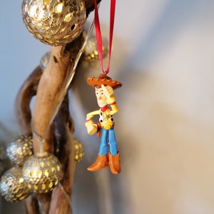 Pu&ograve; includere: Un filo rosso pende da un ramo con una statuetta di plastica di Woody di Toy Story. Woody indossa il suo cappello marrone distintivo, una camicia gialla, jeans blu e stivali marroni. Sta tenendo la mano destra sulla fronte, come se stesse salutando.