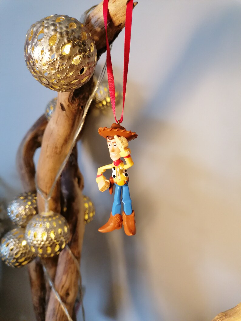 Pu&ograve; includere: Un filo rosso pende da un ramo con una statuetta di plastica di Woody di Toy Story. Woody indossa il suo cappello marrone distintivo, una camicia gialla, jeans blu e stivali marroni. Sta tenendo la mano destra sulla fronte, come se stesse salutando.