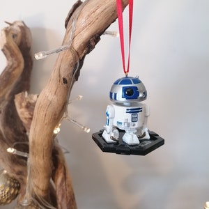 May include: A white and blue R2-D2 Star Wars character ornament hanging from a red ribbon on a branch with white lights.