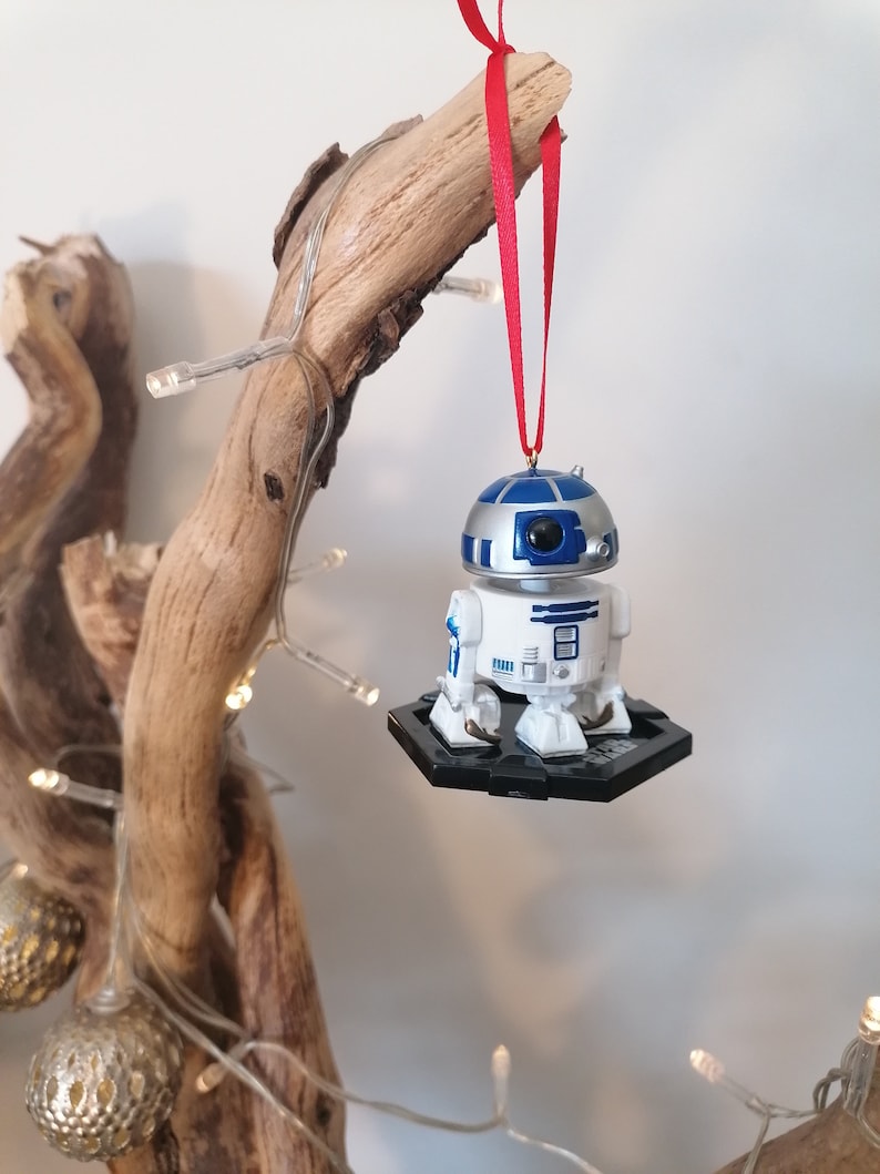 May include: A white and blue R2-D2 Star Wars character ornament hanging from a red ribbon on a branch with white lights.