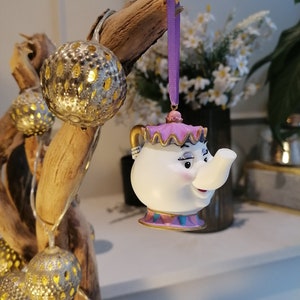 May include: A white porcelain teapot ornament with a purple and pink lid and a smiling face. The teapot is hanging from a purple ribbon.