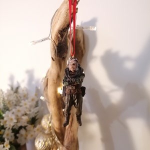 K&ouml;nnte beinhalten: Detaillierter Weihnachtsschmuck, der eine Figur in Milit&auml;runiform mit Brille und ernstem Gesichtsausdruck darstellt. Der Schmuck wird von einem roten Band gehalten. Die Figur ist braun, gr&uuml;n und schwarz. Der Hintergrund ist ein St&uuml;ck Treibholz.