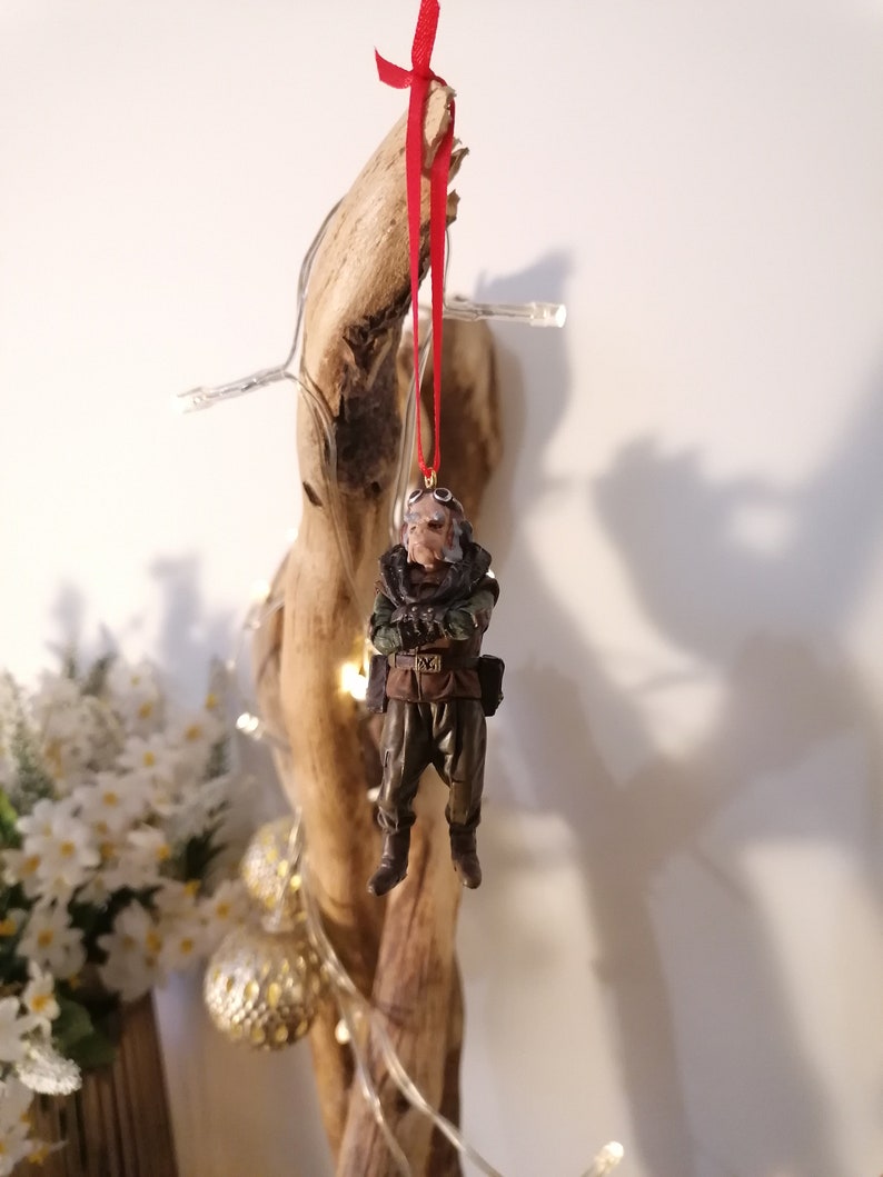 K&ouml;nnte beinhalten: Detaillierter Weihnachtsschmuck, der eine Figur in Milit&auml;runiform mit Brille und ernstem Gesichtsausdruck darstellt. Der Schmuck wird von einem roten Band gehalten. Die Figur ist braun, gr&uuml;n und schwarz. Der Hintergrund ist ein St&uuml;ck Treibholz.