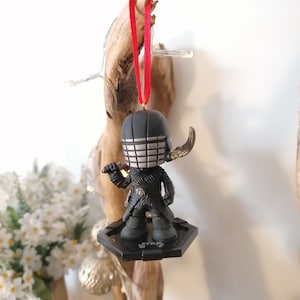 May include: Black and silver Star Wars ornament featuring a character with a helmet and a lightsaber. The ornament is hanging from a red ribbon.