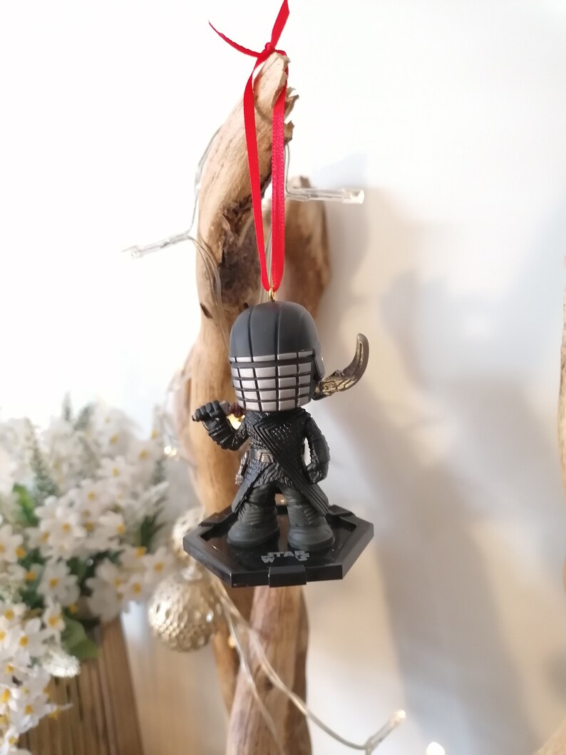 May include: Black and silver Star Wars ornament featuring a character with a helmet and a lightsaber. The ornament is hanging from a red ribbon.