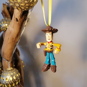 May include: A Woody action figure ornament, featuring a yellow shirt, blue pants, and brown boots, hangs from a yellow ribbon. The ornament is suspended in front of a wooden branch with decorative gold sphere lights.