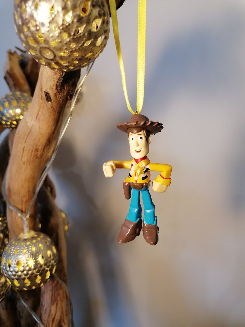 May include: A Woody action figure ornament, featuring a yellow shirt, blue pants, and brown boots, hangs from a yellow ribbon. The ornament is suspended in front of a wooden branch with decorative gold sphere lights.