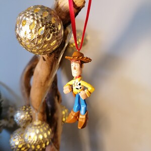 May include: A red and white string hangs from a piece of driftwood with a gold ornament. A small plastic figurine of Woody from Toy Story is hanging from the string.