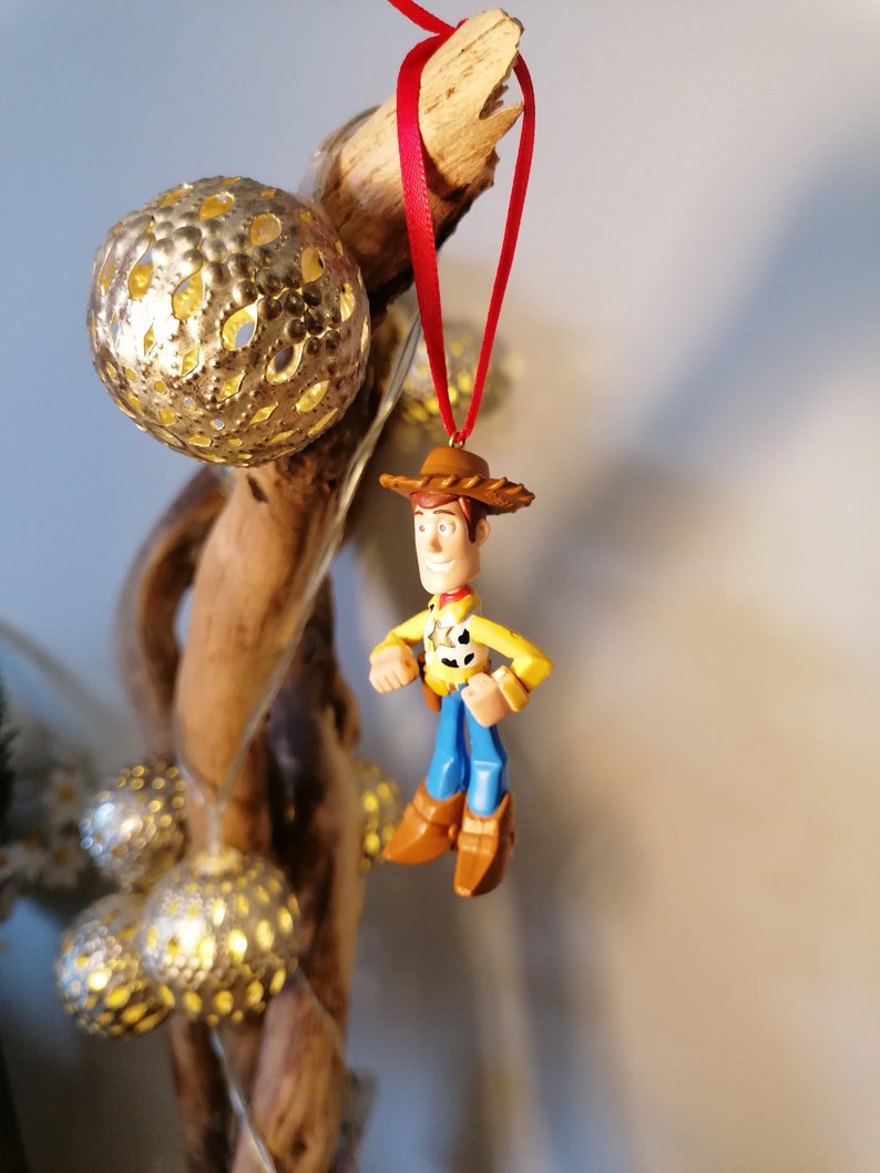 May include: A red and white string hangs from a piece of driftwood with a gold ornament. A small plastic figurine of Woody from Toy Story is hanging from the string.