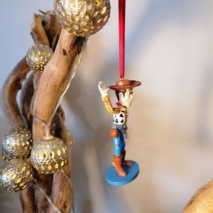 May include: A red string hangs from a branch with a small, blue-based figurine of Woody from Toy Story. Woody is wearing his signature cowboy hat, vest, and jeans. He is waving with his right hand.