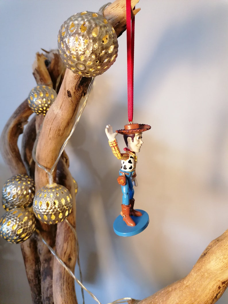 May include: A red string hangs from a branch with a small, blue-based figurine of Woody from Toy Story. Woody is wearing his signature cowboy hat, vest, and jeans. He is waving with his right hand.
