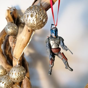 May include: A blue and gray Star Wars Boba Fett action figure ornament hanging from a branch with a red ribbon. The ornament is decorated with silver and gold balls.