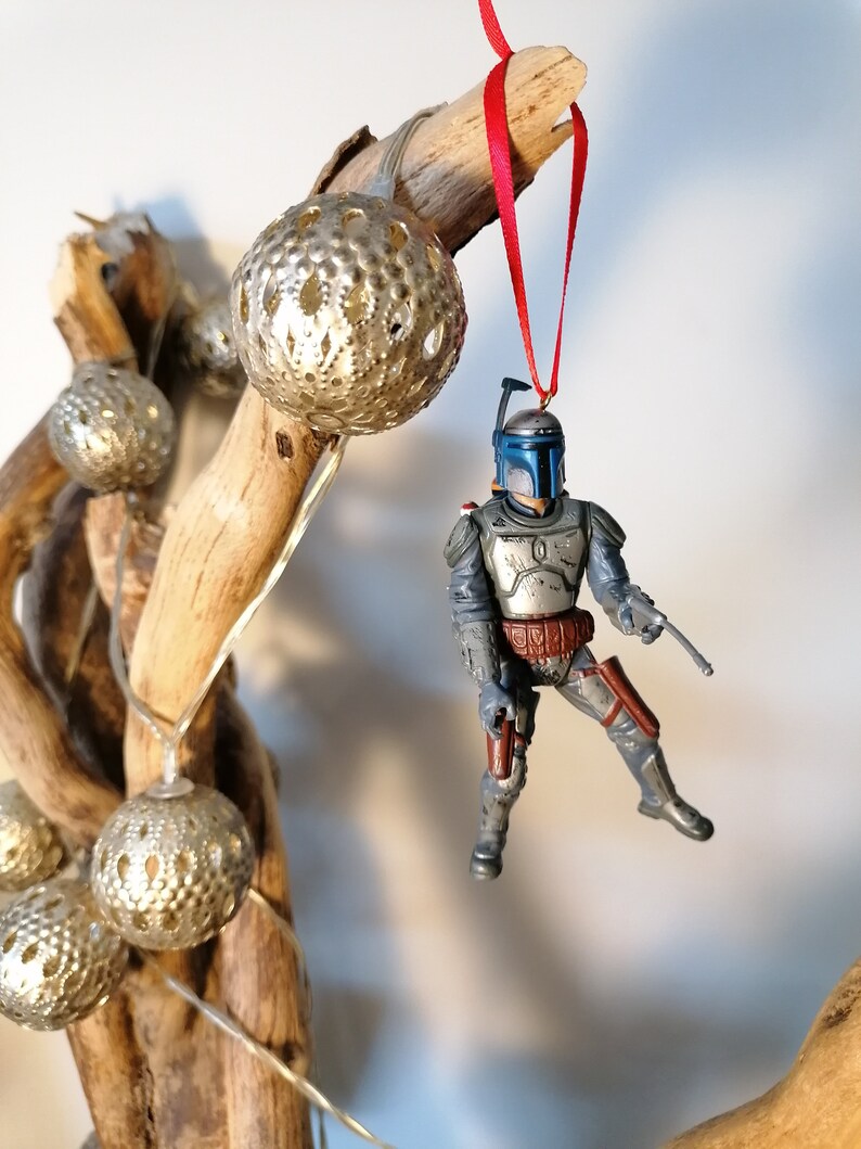 May include: A blue and gray Star Wars Boba Fett action figure ornament hanging from a branch with a red ribbon. The ornament is decorated with silver and gold balls.