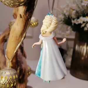 May include: A blue and white dress ornament of a Disney princess with blonde hair. The ornament is hanging from a silver string.