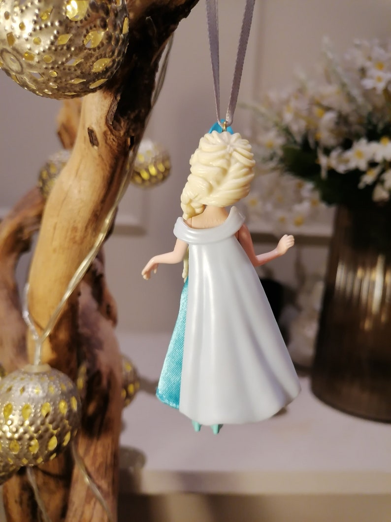 May include: A blue and white dress ornament of a Disney princess with blonde hair. The ornament is hanging from a silver string.