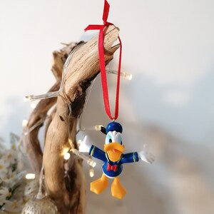 May include: A blue and yellow Donald Duck Christmas ornament with a red bow hanging from a branch with white Christmas lights.