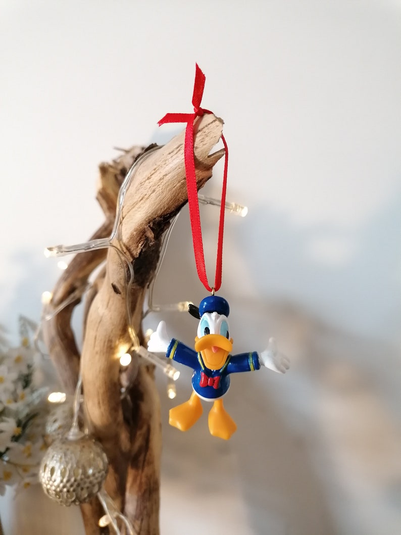 May include: A blue and yellow Donald Duck Christmas ornament with a red bow hanging from a branch with white Christmas lights.