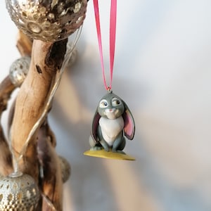 May include: A gray rabbit ornament with pink ears and a white chest, hanging from a pink ribbon. The ornament is sitting on a yellow base.