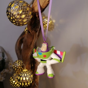 May include: A purple and green Buzz Lightyear toy ornament hanging from a branch with a purple ribbon. The ornament is made of plastic and has a detailed design.