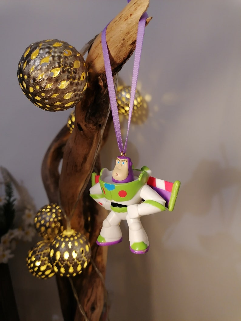May include: A purple and green Buzz Lightyear toy ornament hanging from a branch with a purple ribbon. The ornament is made of plastic and has a detailed design.
