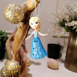 May include: A blue and white plastic figurine of a girl with blonde hair wearing a blue dress with snowflake designs. The figurine is hanging from a blue ribbon on a branch.