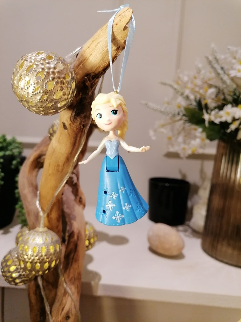 May include: A blue and white plastic figurine of a girl with blonde hair wearing a blue dress with snowflake designs. The figurine is hanging from a blue ribbon on a branch.
