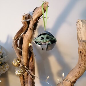 May include: A green and gray Christmas ornament featuring a character from the Star Wars franchise, The Mandalorian. The ornament is hanging from a branch of a driftwood tree decorated with white fairy lights.