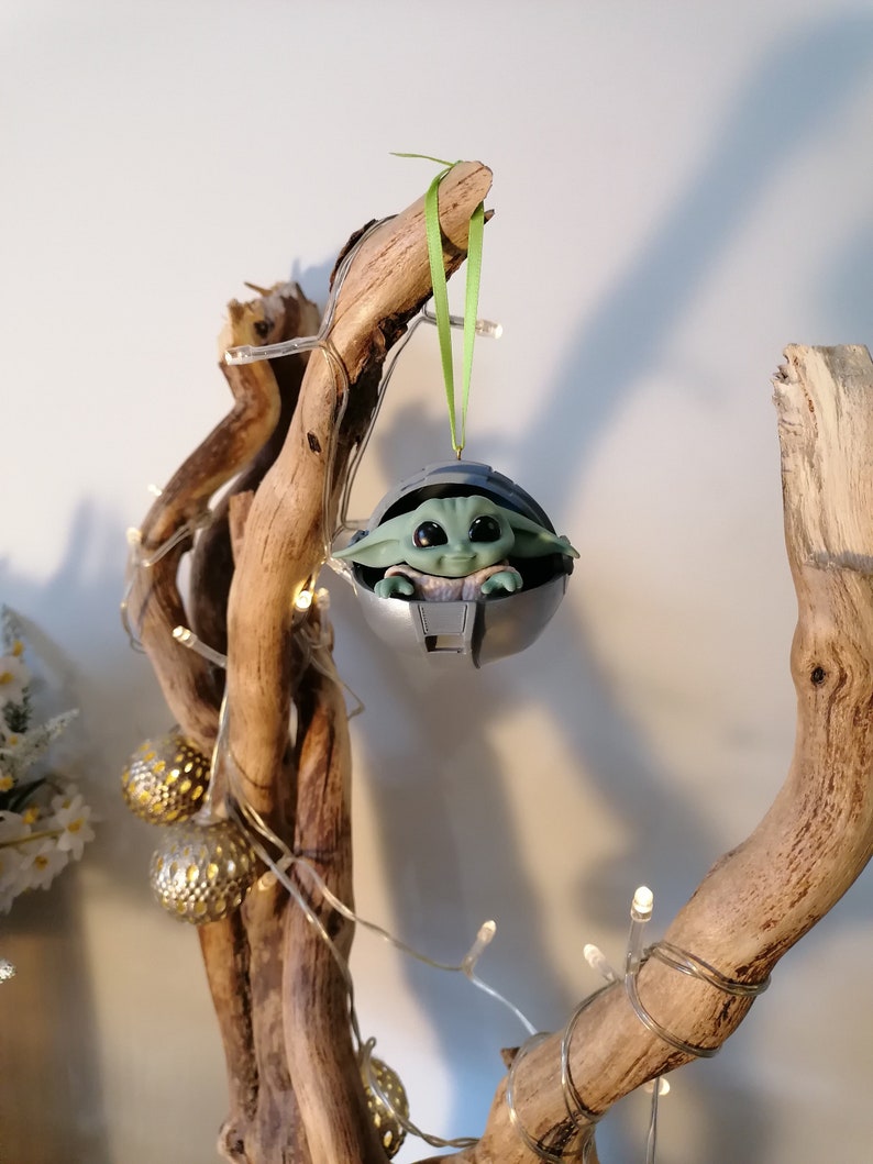 May include: A green and gray Christmas ornament featuring a character from the Star Wars franchise, The Mandalorian. The ornament is hanging from a branch of a driftwood tree decorated with white fairy lights.