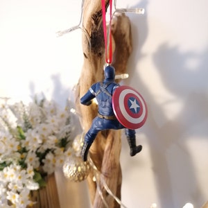 May include: A blue and red Captain America Christmas ornament hanging from a branch. The ornament features a detailed figure of Captain America holding his shield.
