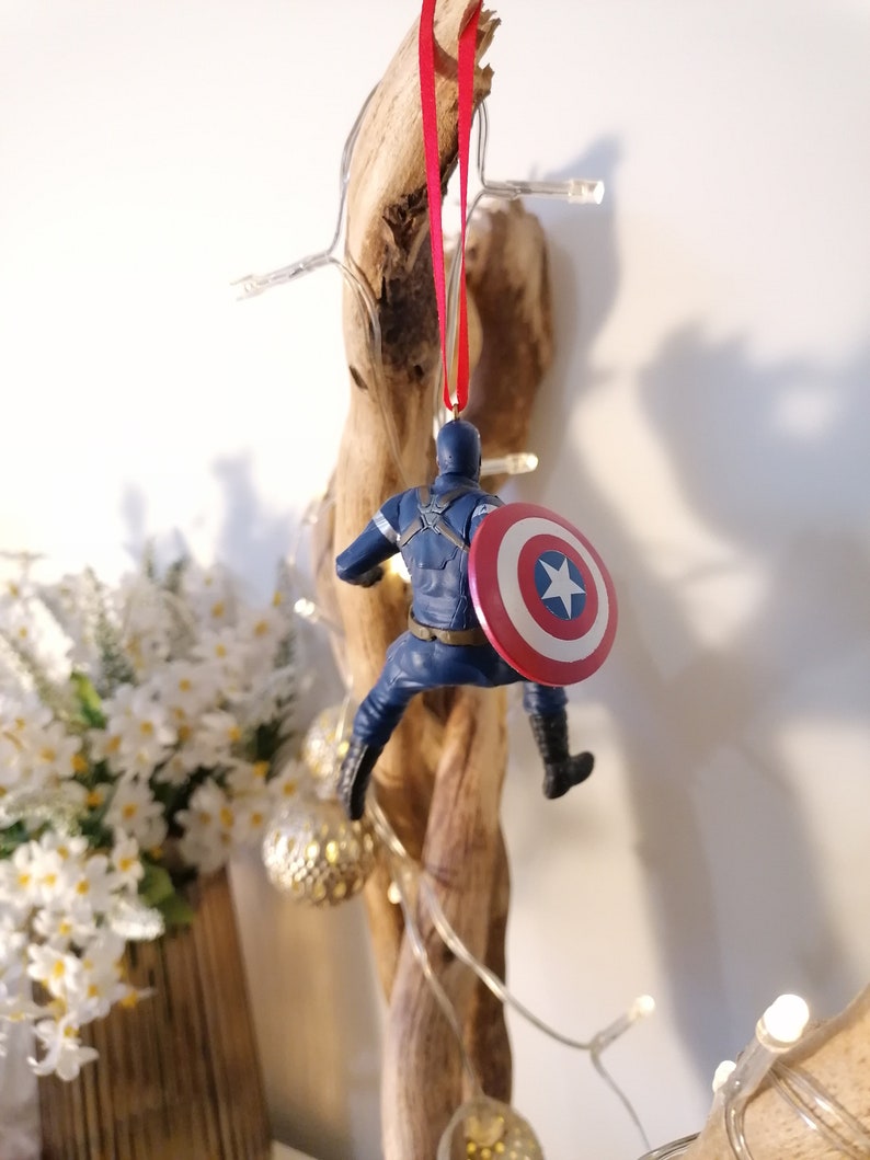May include: A blue and red Captain America Christmas ornament hanging from a branch. The ornament features a detailed figure of Captain America holding his shield.