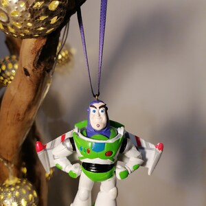 Pu&ograve; includere: Un ornamento di giocattolo in plastica verde e bianco di Buzz Lightyear del franchise Toy Story. L'ornamento &egrave; appeso a un nastro viola e presenta la tuta da ranger spaziale di Buzz Lightyear, il suo casco e le sue ali.