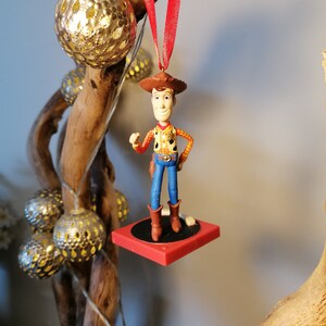 Puede incluir: Un adorno navide&ntilde;o rojo y blanco con una figura de pl&aacute;stico de Woody de la pel&iacute;cula Toy Story. Woody lleva su caracter&iacute;stico sombrero de vaquero, chaleco y pantalones vaqueros. Est&aacute; de pie sobre una plataforma roja con una base negra.