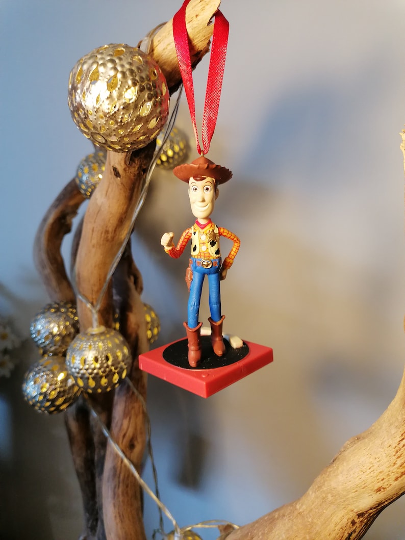 Puede incluir: Un adorno navide&ntilde;o rojo y blanco con una figura de pl&aacute;stico de Woody de la pel&iacute;cula Toy Story. Woody lleva su caracter&iacute;stico sombrero de vaquero, chaleco y pantalones vaqueros. Est&aacute; de pie sobre una plataforma roja con una base negra.