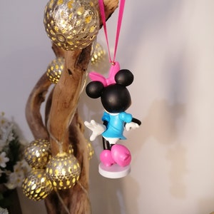 May include: A pink and blue Minnie Mouse ornament with a pink bow hanging from a pink ribbon. The ornament is hanging from a branch with string lights.