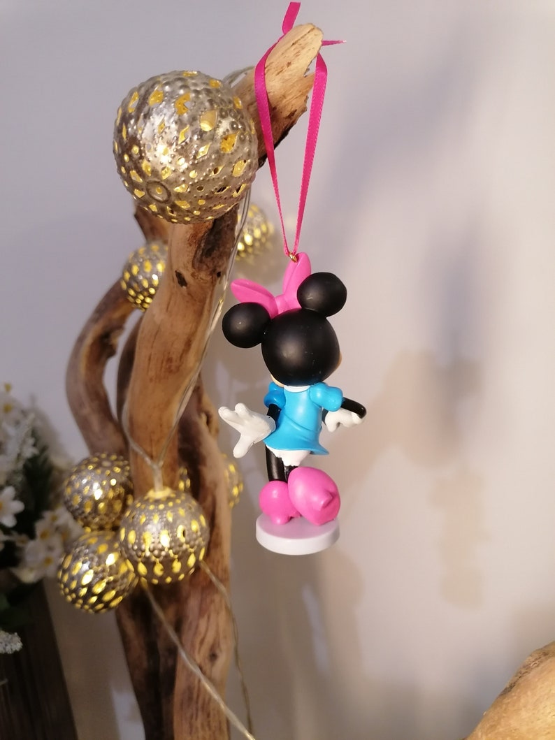May include: A pink and blue Minnie Mouse ornament with a pink bow hanging from a pink ribbon. The ornament is hanging from a branch with string lights.