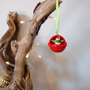 Pu&ograve; includere: Un ornamento natalizio rosso con una figura verde che indossa un cappello da Babbo Natale all'interno. L'ornamento &egrave; appeso a un nastro verde su un ramo di legno di deriva con luci bianche.