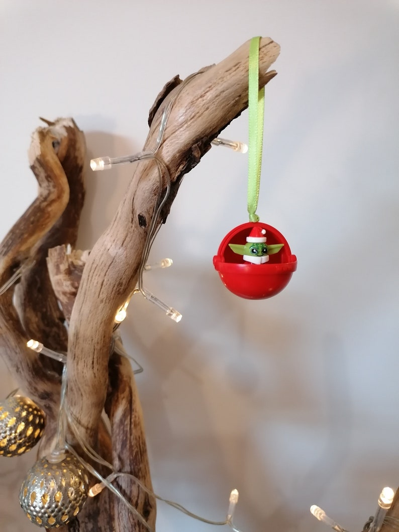 Pu&ograve; includere: Un ornamento natalizio rosso con una figura verde che indossa un cappello da Babbo Natale all'interno. L'ornamento &egrave; appeso a un nastro verde su un ramo di legno di deriva con luci bianche.
