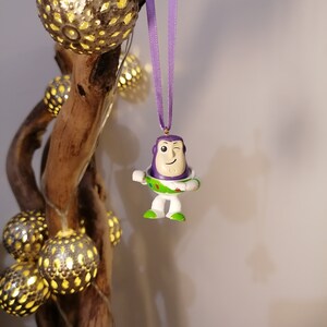 May include: Purple and white plastic ornament of Buzz Lightyear from the Toy Story franchise, with a winking face, hanging from a purple ribbon on a branch with string lights.