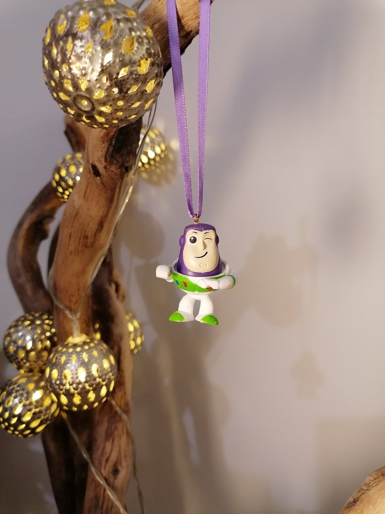 May include: Purple and white plastic ornament of Buzz Lightyear from the Toy Story franchise, with a winking face, hanging from a purple ribbon on a branch with string lights.