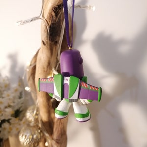 K&ouml;nnte beinhalten: Lila und gr&uuml;ner Buzz Lightyear Weihnachtsbaumschmuck mit Fl&uuml;geln und wei&szlig;em K&ouml;rper. Der Anh&auml;nger h&auml;ngt an einem lila Band.