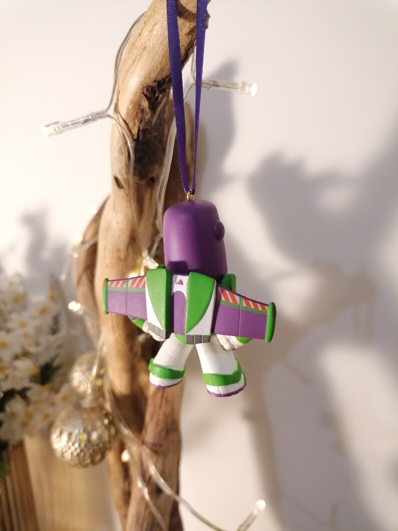 K&ouml;nnte beinhalten: Lila und gr&uuml;ner Buzz Lightyear Weihnachtsbaumschmuck mit Fl&uuml;geln und wei&szlig;em K&ouml;rper. Der Anh&auml;nger h&auml;ngt an einem lila Band.