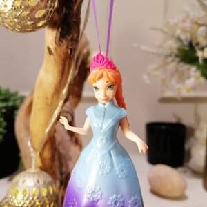 K&ouml;nnte beinhalten: Eine Plastikpuppe mit orangefarbenem Haar und einer rosa Krone, die ein lila-blaues Kleid mit Schneeflocken-Details tr&auml;gt. Die Puppe h&auml;ngt an einer lila Schnur.