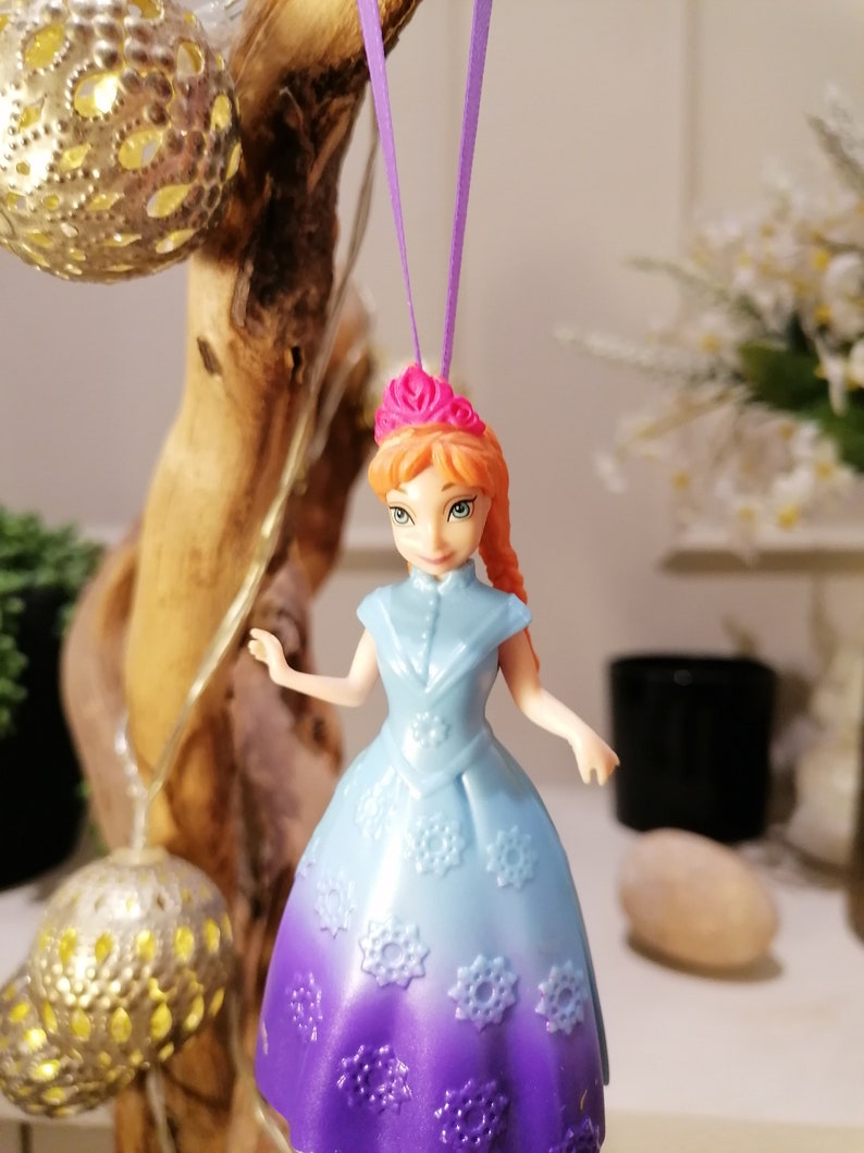 K&ouml;nnte beinhalten: Eine Plastikpuppe mit orangefarbenem Haar und einer rosa Krone, die ein lila-blaues Kleid mit Schneeflocken-Details tr&auml;gt. Die Puppe h&auml;ngt an einer lila Schnur.