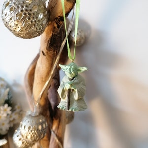 May include: A green Yoda ornament hanging from a branch with a green ribbon. The ornament is made of plastic and has a detailed design.