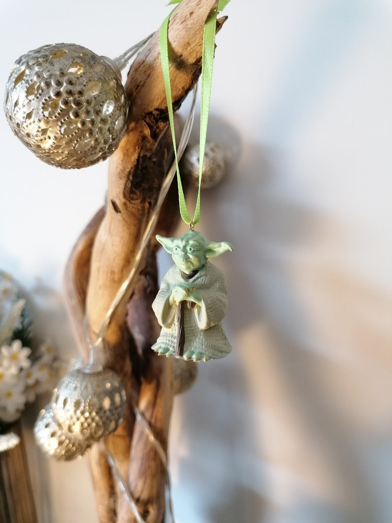 May include: A green Yoda ornament hanging from a branch with a green ribbon. The ornament is made of plastic and has a detailed design.