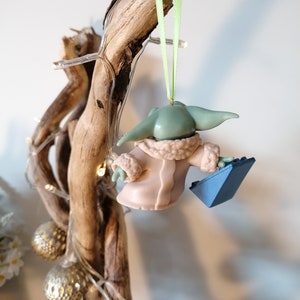 May include: A green and pink Christmas ornament of a character from the Star Wars franchise, holding a blue object. The ornament is hanging from a green ribbon on a branch.