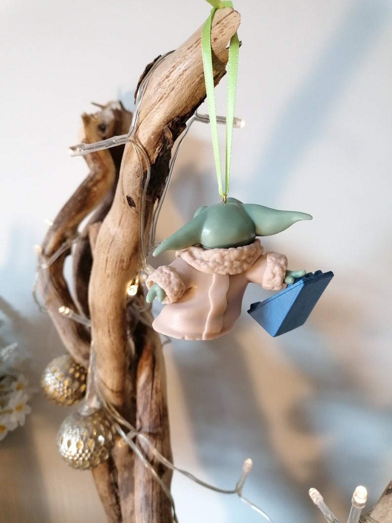 May include: A green and pink Christmas ornament of a character from the Star Wars franchise, holding a blue object. The ornament is hanging from a green ribbon on a branch.