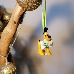 May include: A yellow Pluto Christmas tree decoration, wearing a black hat and holding a bone-shaped sign with "London New York" on it. It hangs from a green ribbon. The background features a wooden branch and fairy lights.